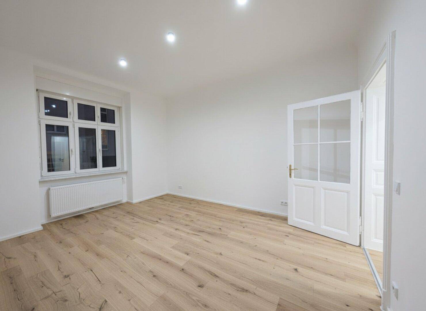 Heller, leerer Raum mit Holzboden und großem Fenster, das viel Tageslicht hereinlässt.