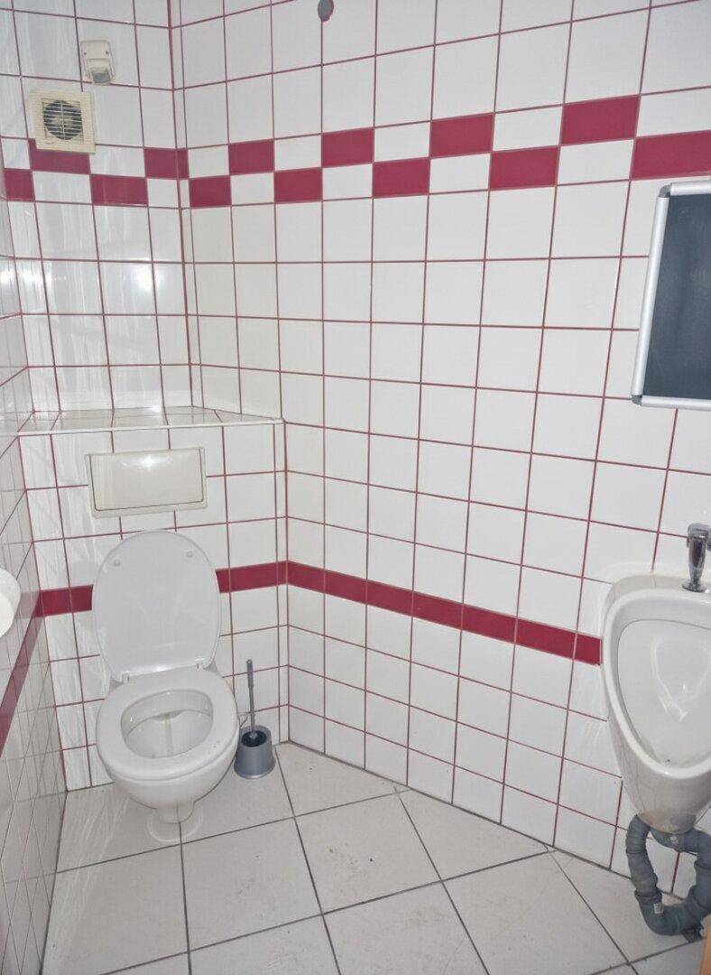 Toilette mit WC, Urinal und weiß-roten Fliesen an den Wänden, sowie einem Abluftventilator.