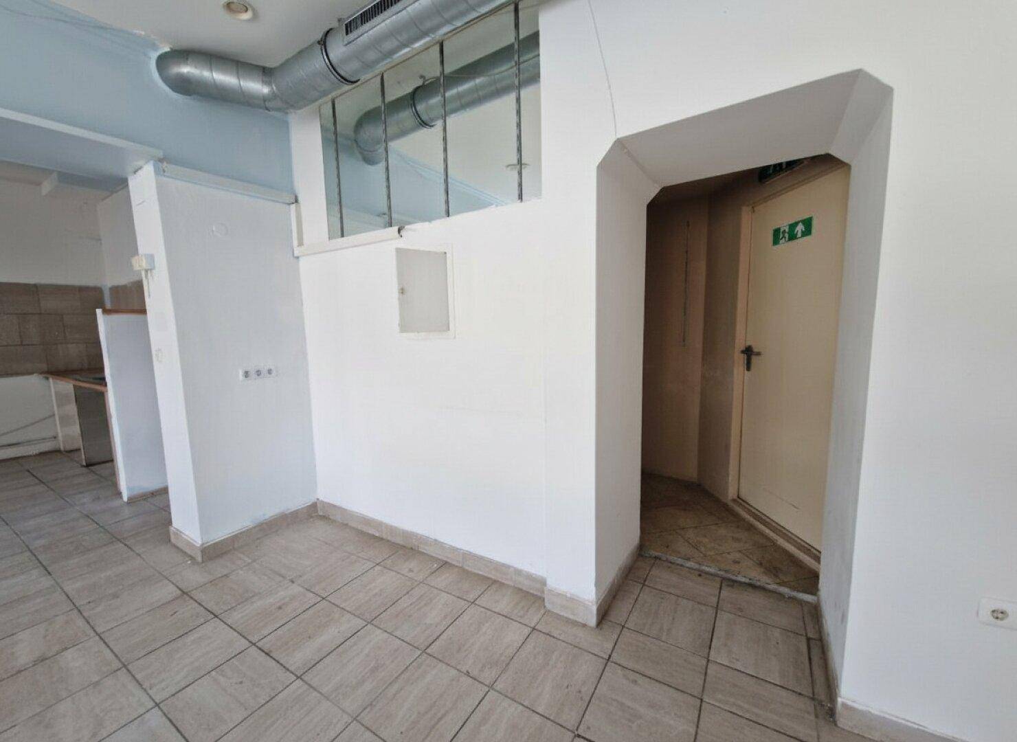 Innenansicht eines Gewerberaums mit Fliesenboden, weiß gestrichenen Wänden und sichtbaren Lüftungsrohren.