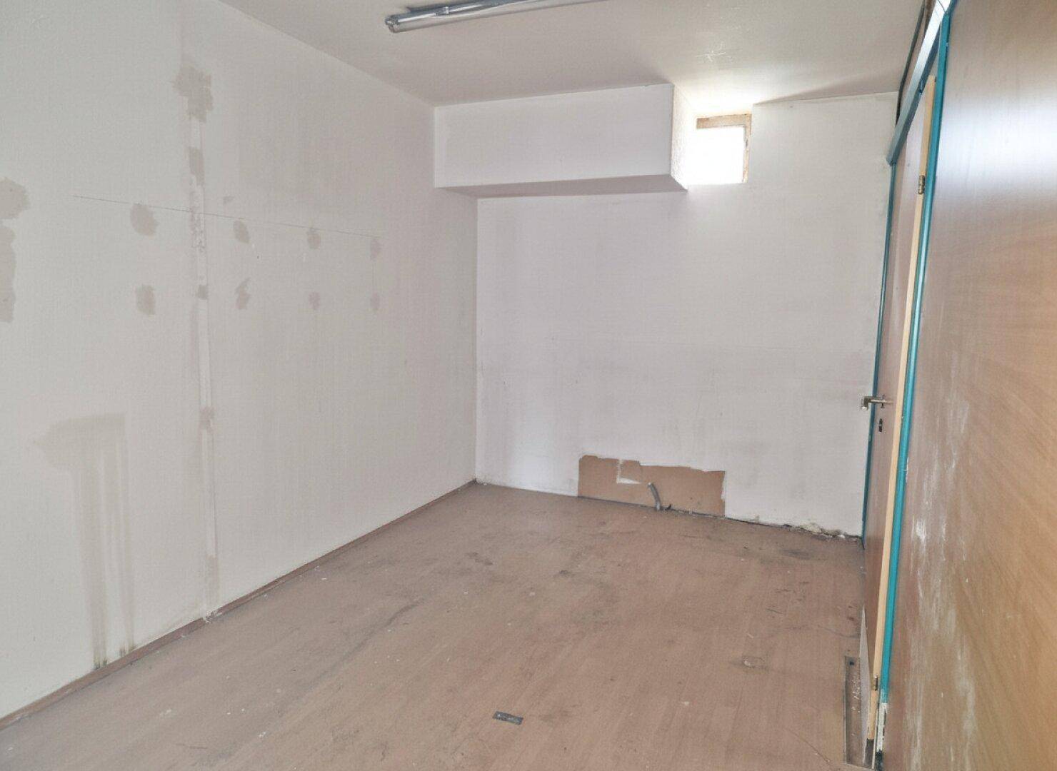 Leerer Raum mit weißen Wänden, Holzboden und einer kleinen Fensteröffnung oben rechts.