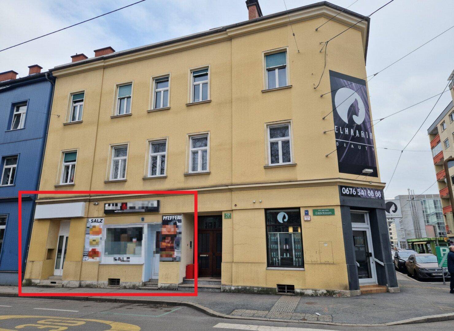 Gelbes Eckgebäude mit mehreren Etagen und einem Ladenlokal im Erdgeschoss an einer Straße.