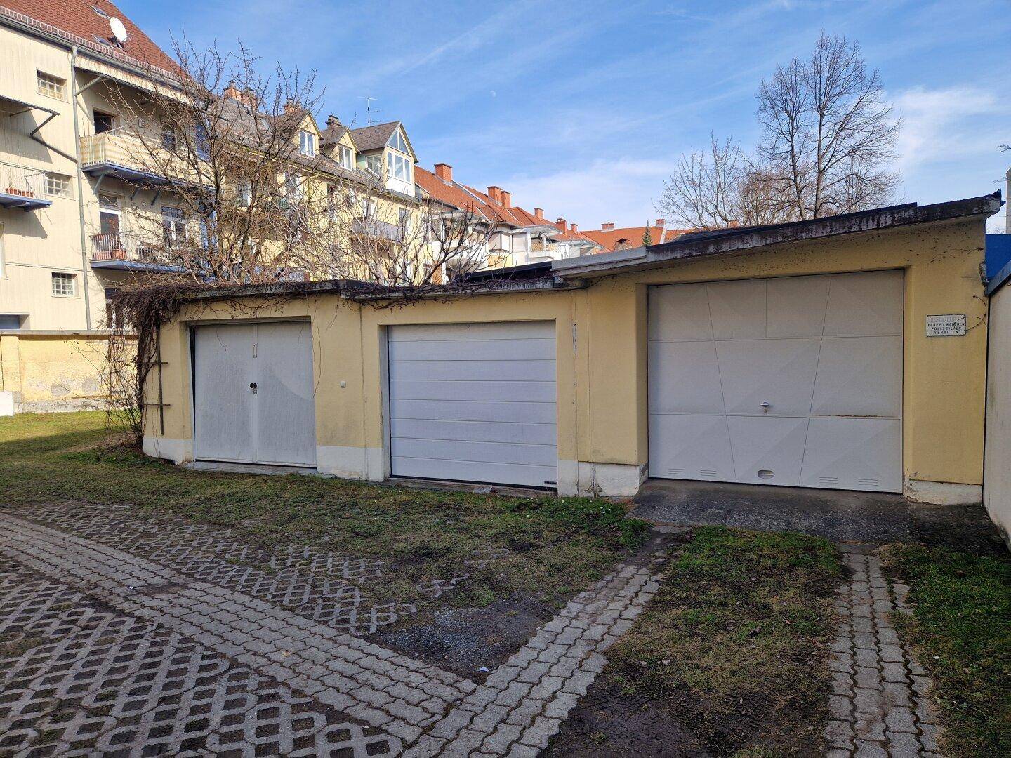 Drei Garagenboxen mit weißen Toren und einem gepflasterten Zufahrtsbereich vor einem Wohngebäude.