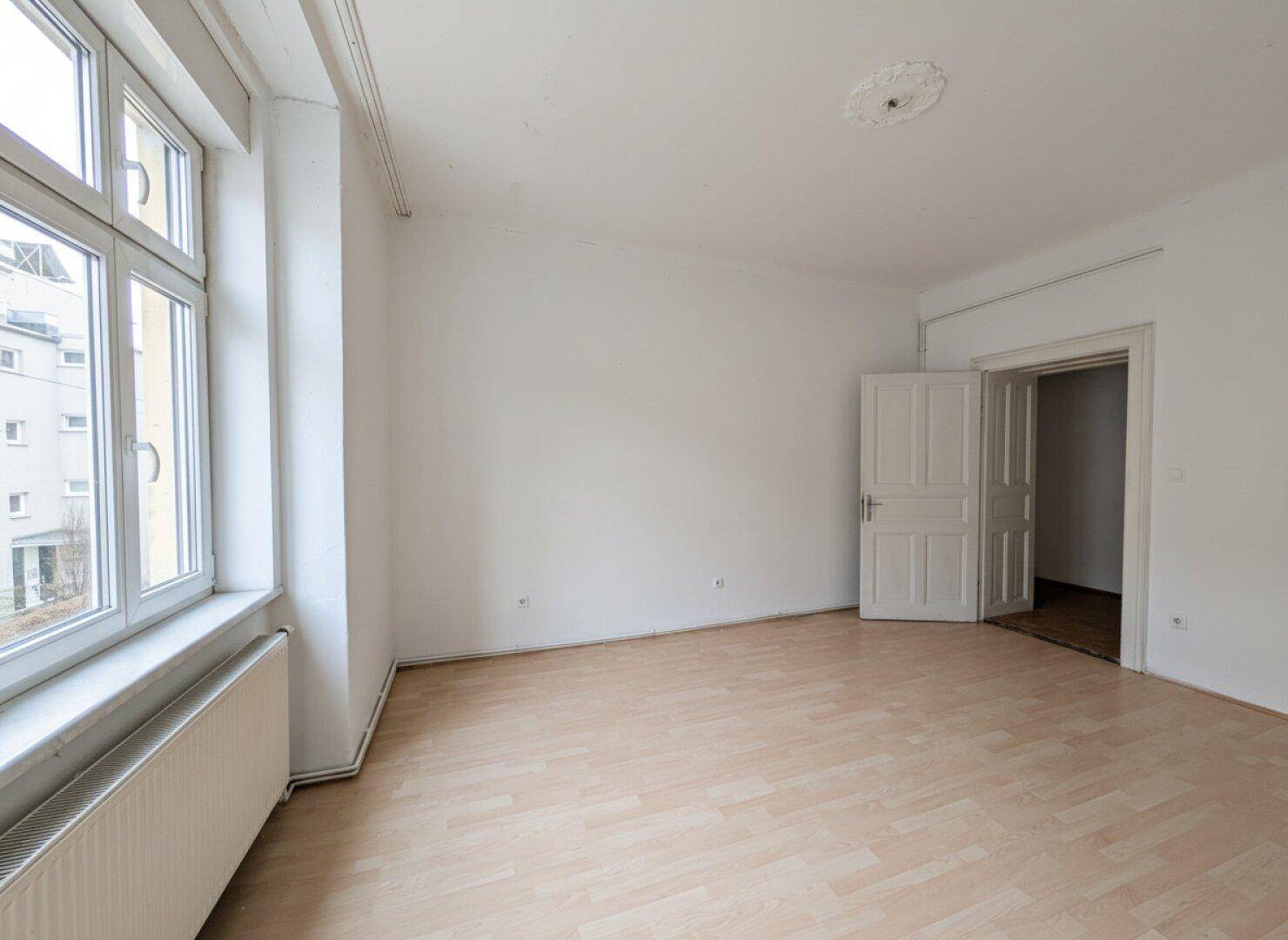Geräumiger, heller Raum mit Laminatboden, Stuckdecke und großem Fenster.