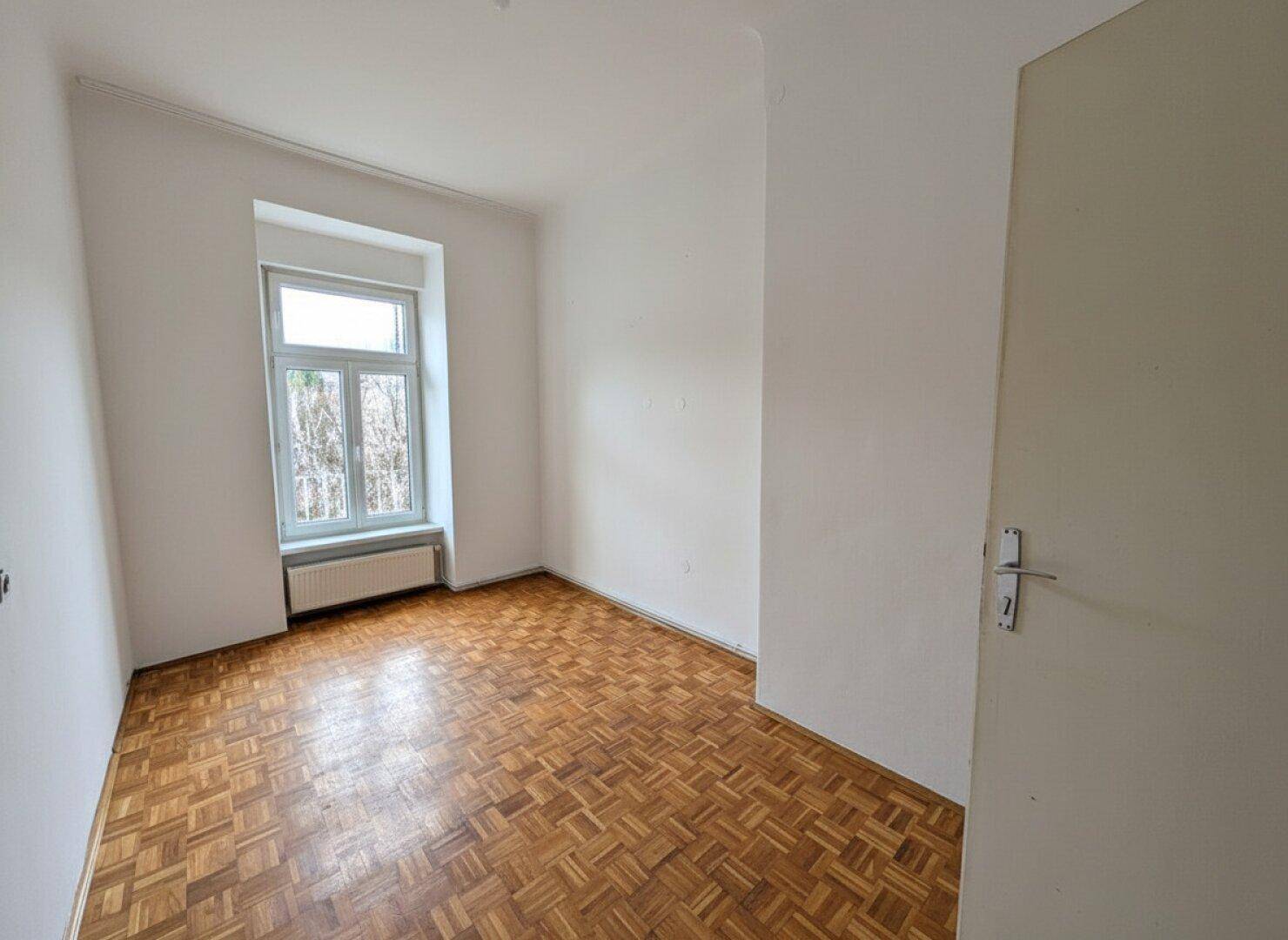 Leerer Raum mit Parkettboden und einem großen Fenster, das viel Licht hereinlässt.