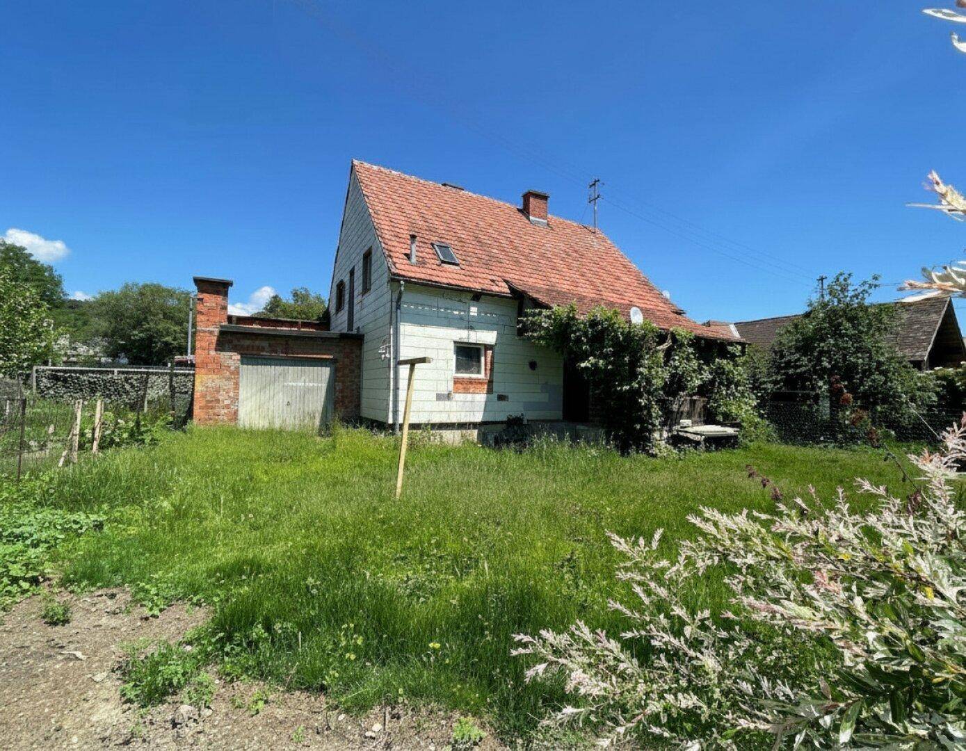 Seitenansicht des Hauses mit verwildertem Garten und viel Grün unter strahlend blauem Himmel.