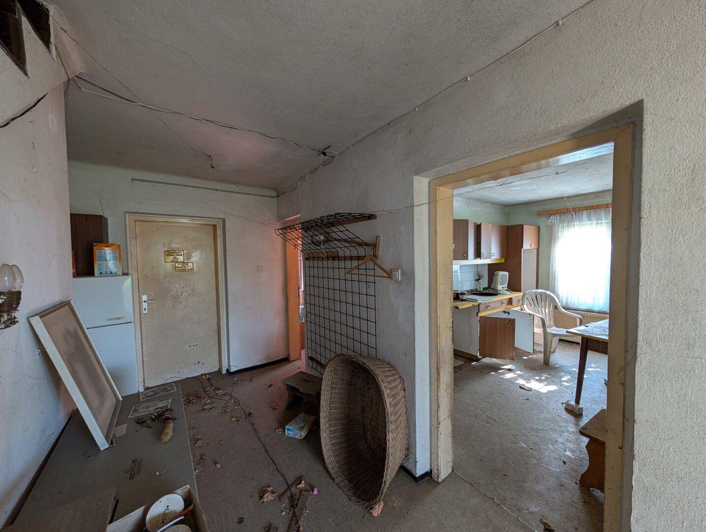 Unaufgeräumter Flur mit alter Einrichtung und Blick in eine angrenzende Küche, die Renovierung benötigt.