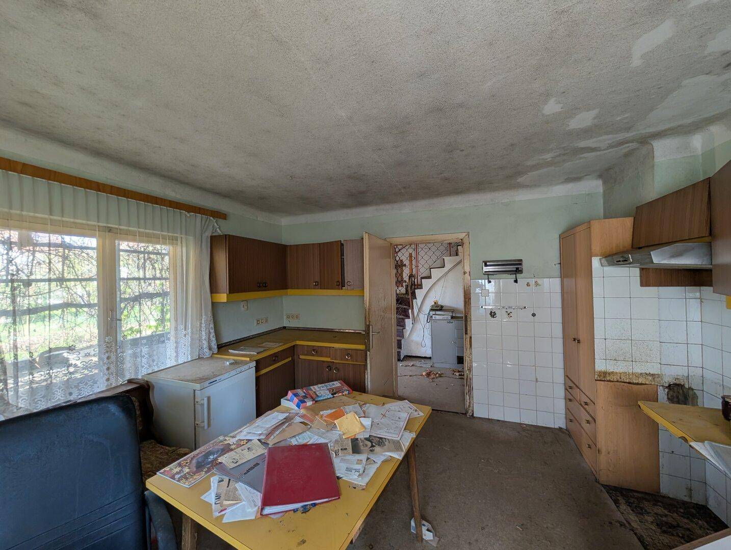 Veraltete Küche mit braunen Holzschränken, Fliesenrückwand und einem Fenster zum Garten.