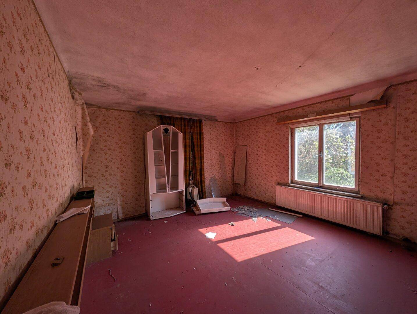 Zimmer mit rotem Teppichboden, floraler Tapete und einem Fenster, das viel Sonnenlicht hereinlässt.