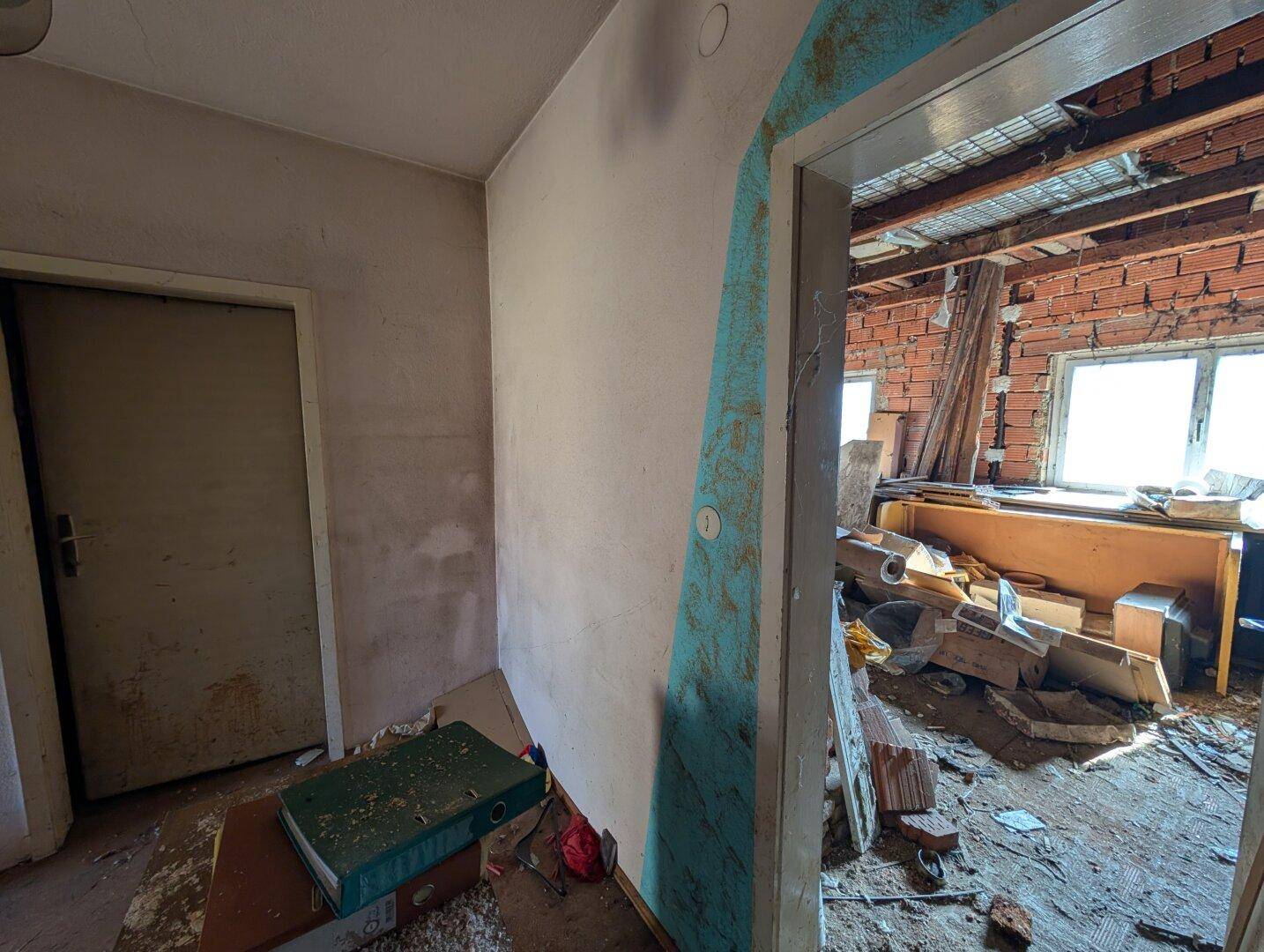 Flur mit Blick in einen Raum, der sich im Rohbau oder Abrisszustand befindet, mit sichtbaren Ziegelwänden.