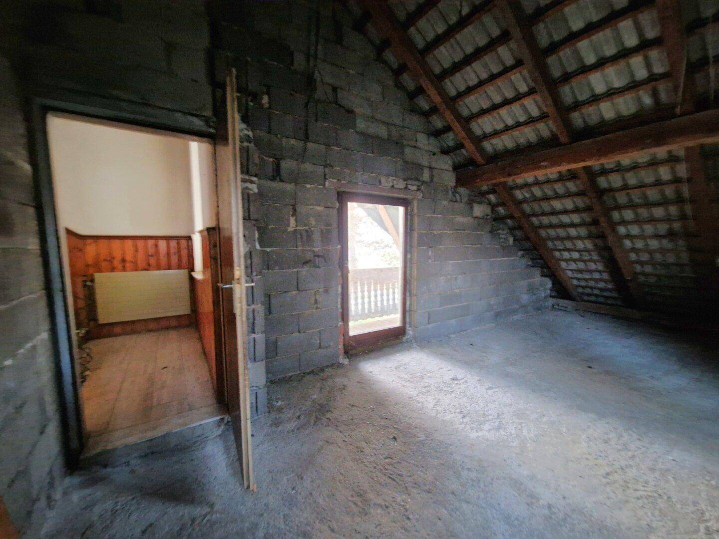 Dachgeschossraum im Rohbau mit schrägen Wänden, Ziegelmauerwerk und Zugang zu einem Balkon.