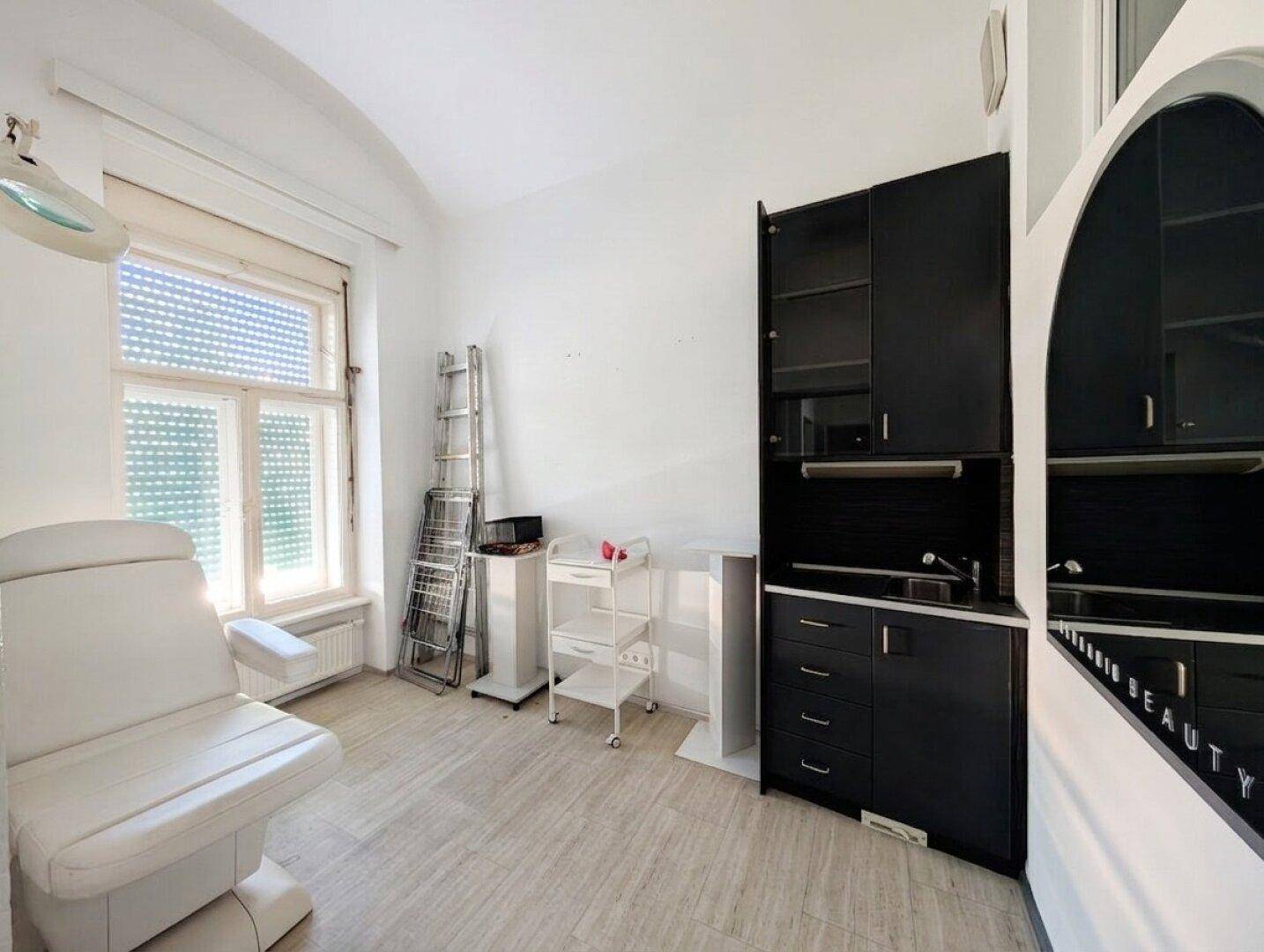 Behandlungsraum mit Liege, Fenster und dunklen Schränken, geeignet für Kosmetik oder Massage mit viel Tageslicht.