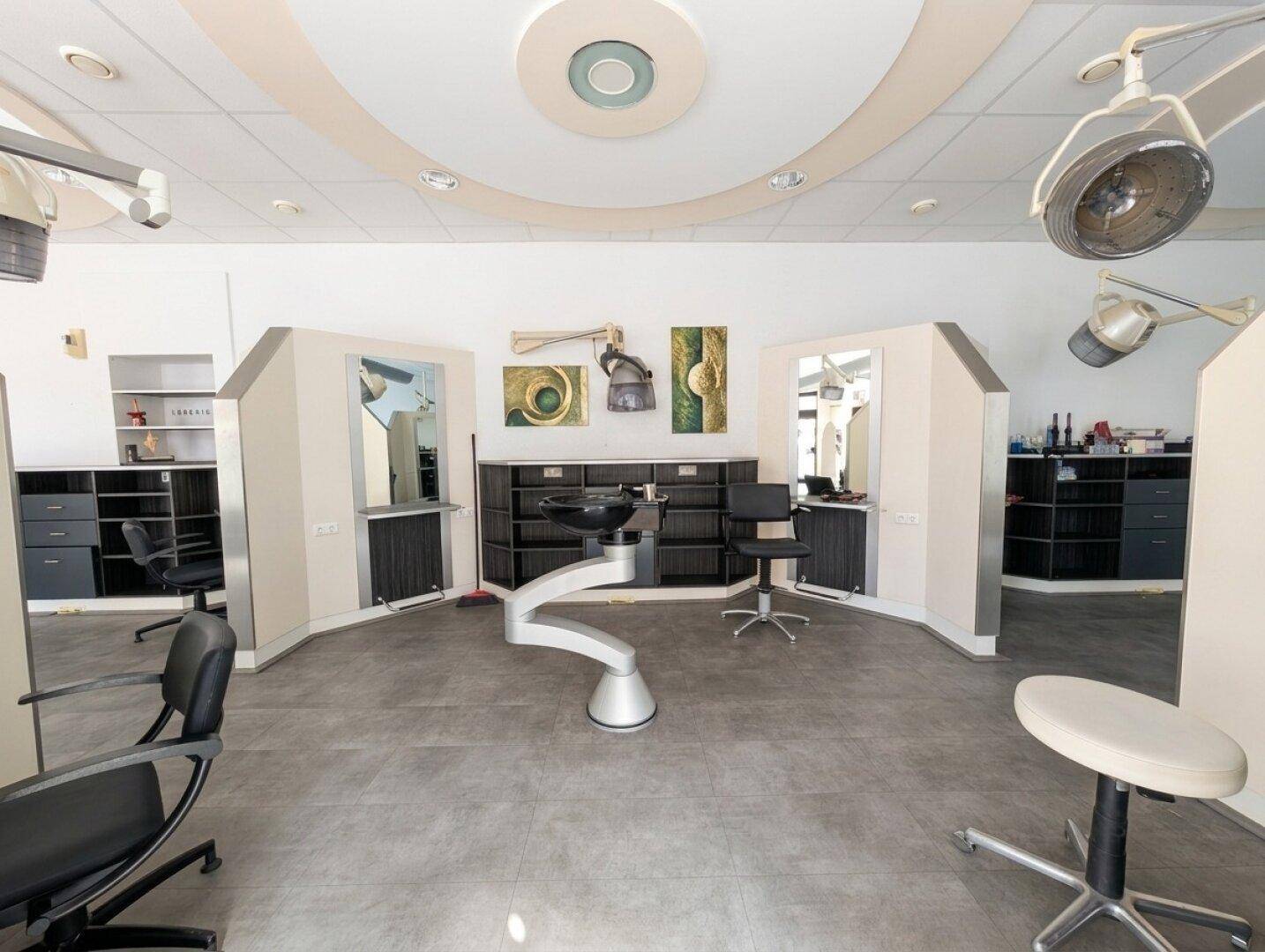 Stylingbereich eines Friseursalons mit mehreren Arbeitsplätzen, Waschbecken und Trockenhauben unter einer hellen Decke.