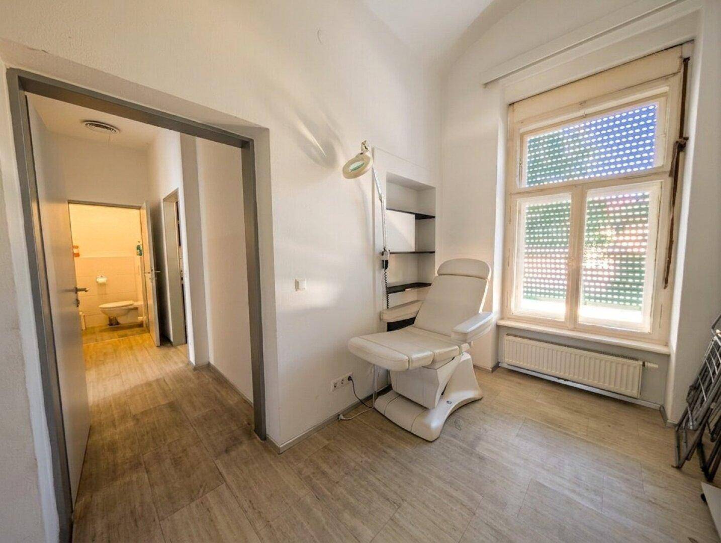 Behandlungsraum mit Liege, Fenster und Zugang zu einem weiteren Raum, heller Boden und weiße Wände.