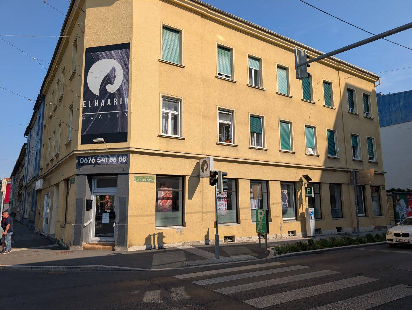 Eckgebäude mit gelber Fassade, Schaufenstern und Werbebanner an einer belebten Straße mit viel Tageslicht.