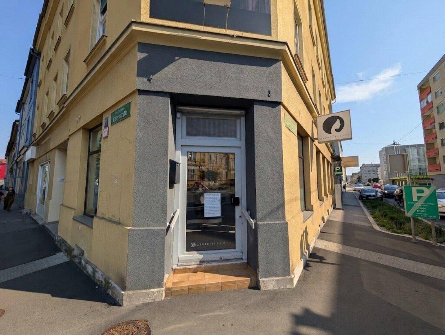 Ecke eines gelben Gebäudes mit Eingangstür und Schaufenstern an einer belebten Straße unter blauem Himmel.