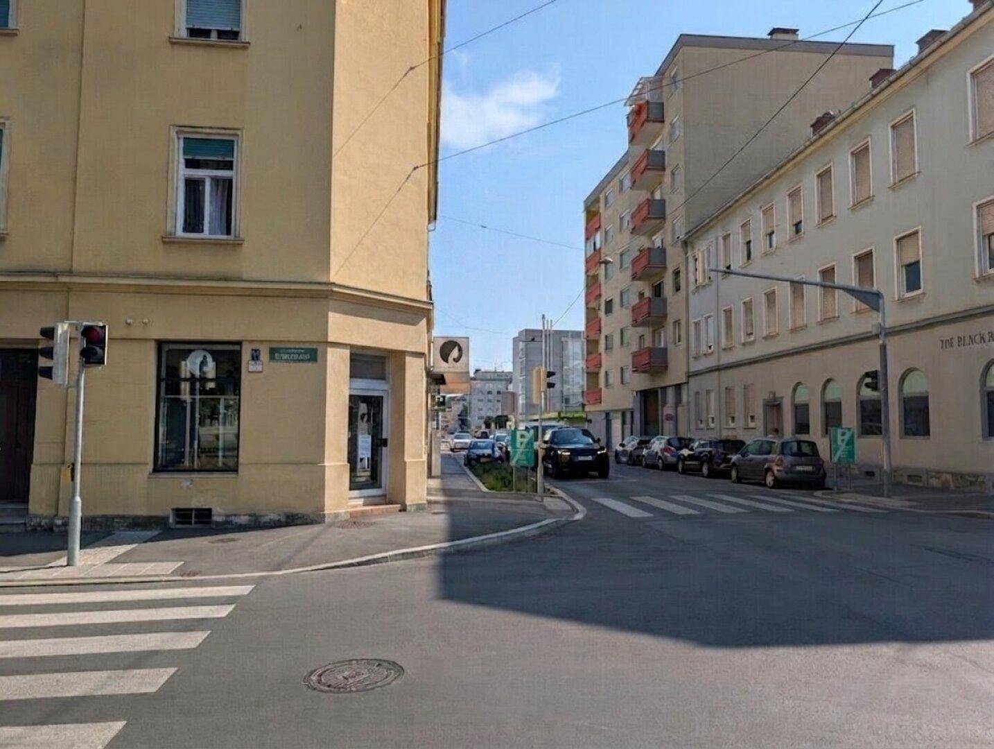 Straßenansicht einer Kreuzung mit Gebäuden, parkenden Autos und Fußgängerüberweg unter blauem Himmel.