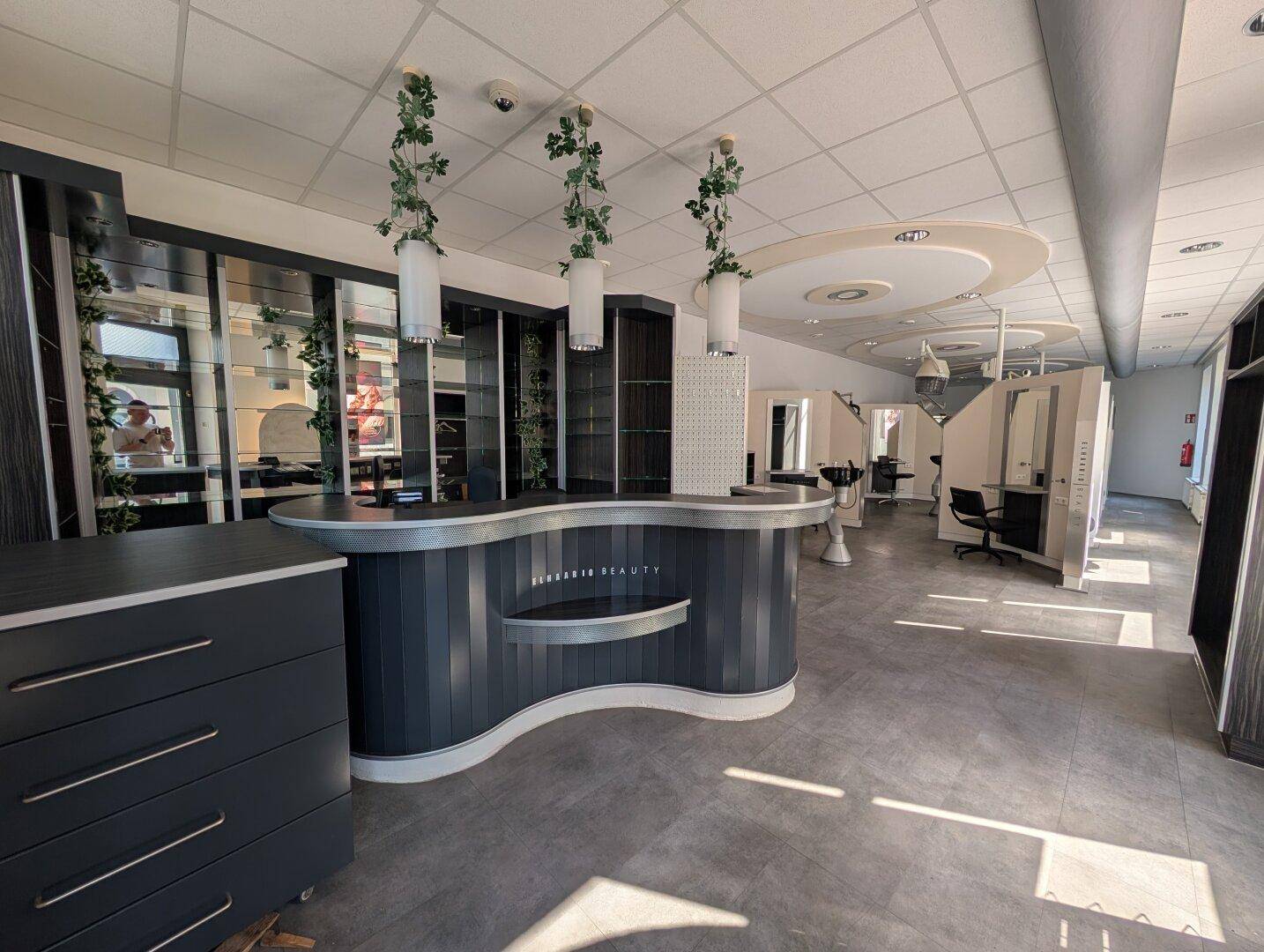 Empfangsbereich eines Friseursalons mit dunkler Theke, Regalen und Blick in den Stylingbereich mit hellen Böden.