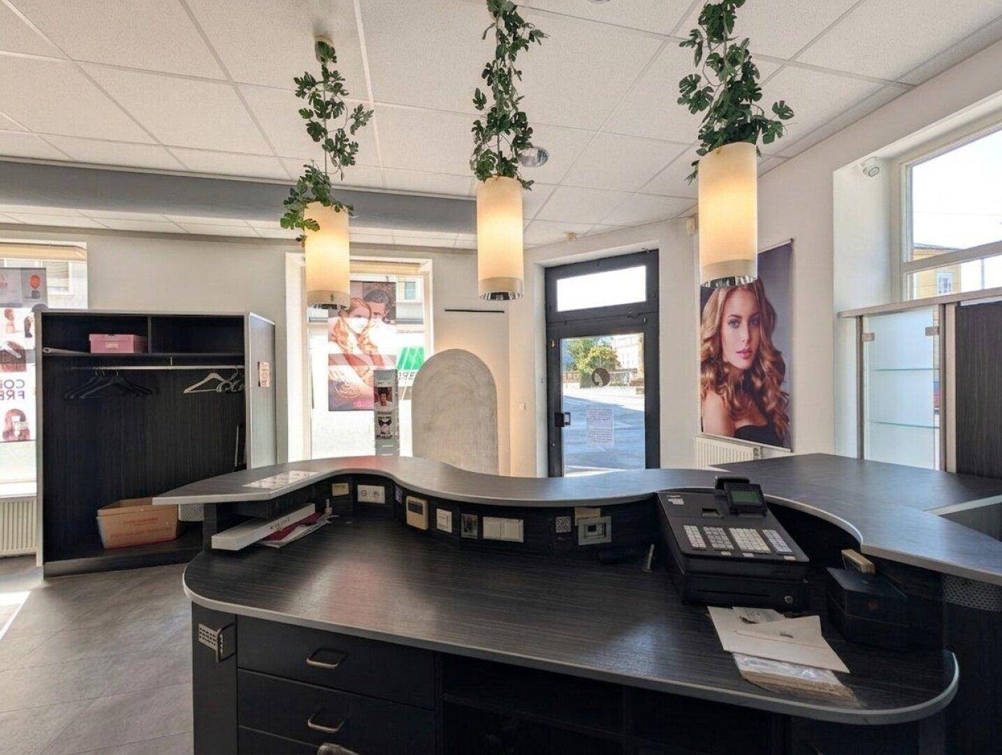 Detailansicht der Empfangstheke eines Salons mit Kasse, Hängeleuchten und dunklen Oberflächen.