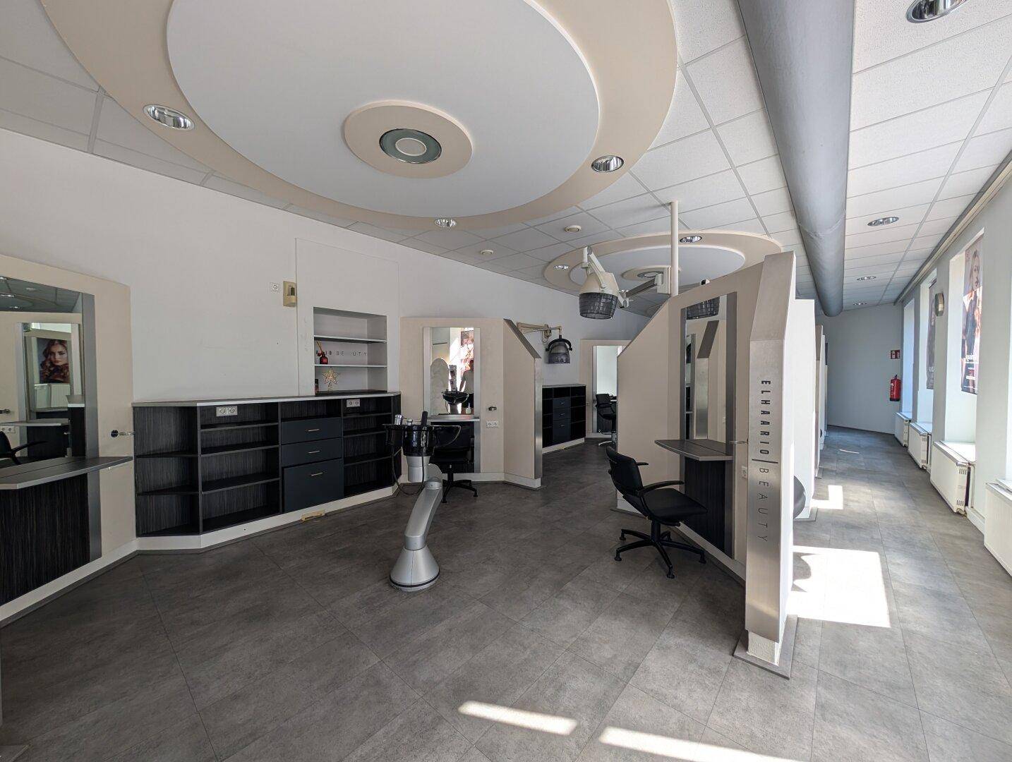 Geräumiger Stylingbereich eines Friseursalons mit mehreren Arbeitsplätzen, Spiegeln und Deckenbeleuchtung.