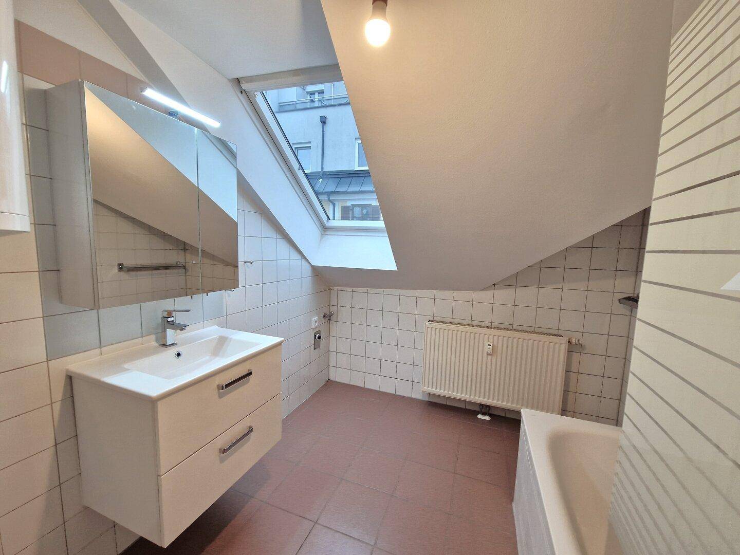 Helles Badezimmer mit Dachfenster, Waschbecken, Spiegelschrank und Fliesenboden.