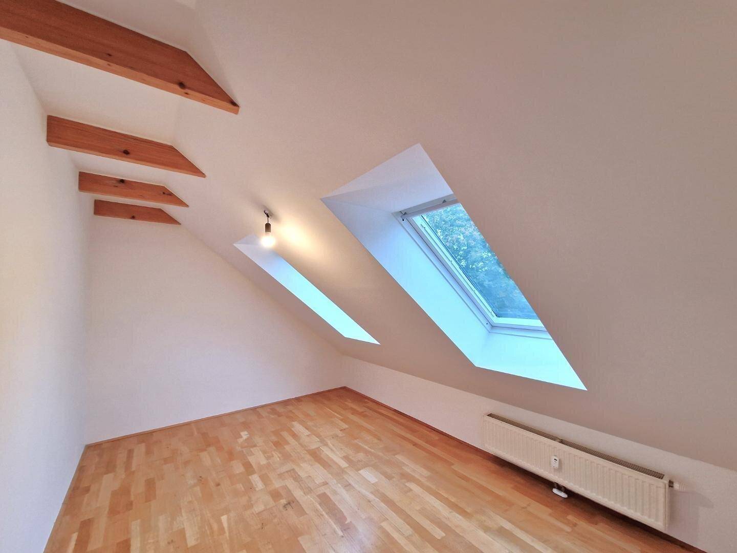 Helles, leeres Schlafzimmer im Dachgeschoss mit zwei Dachfenstern und Holzbalken.
