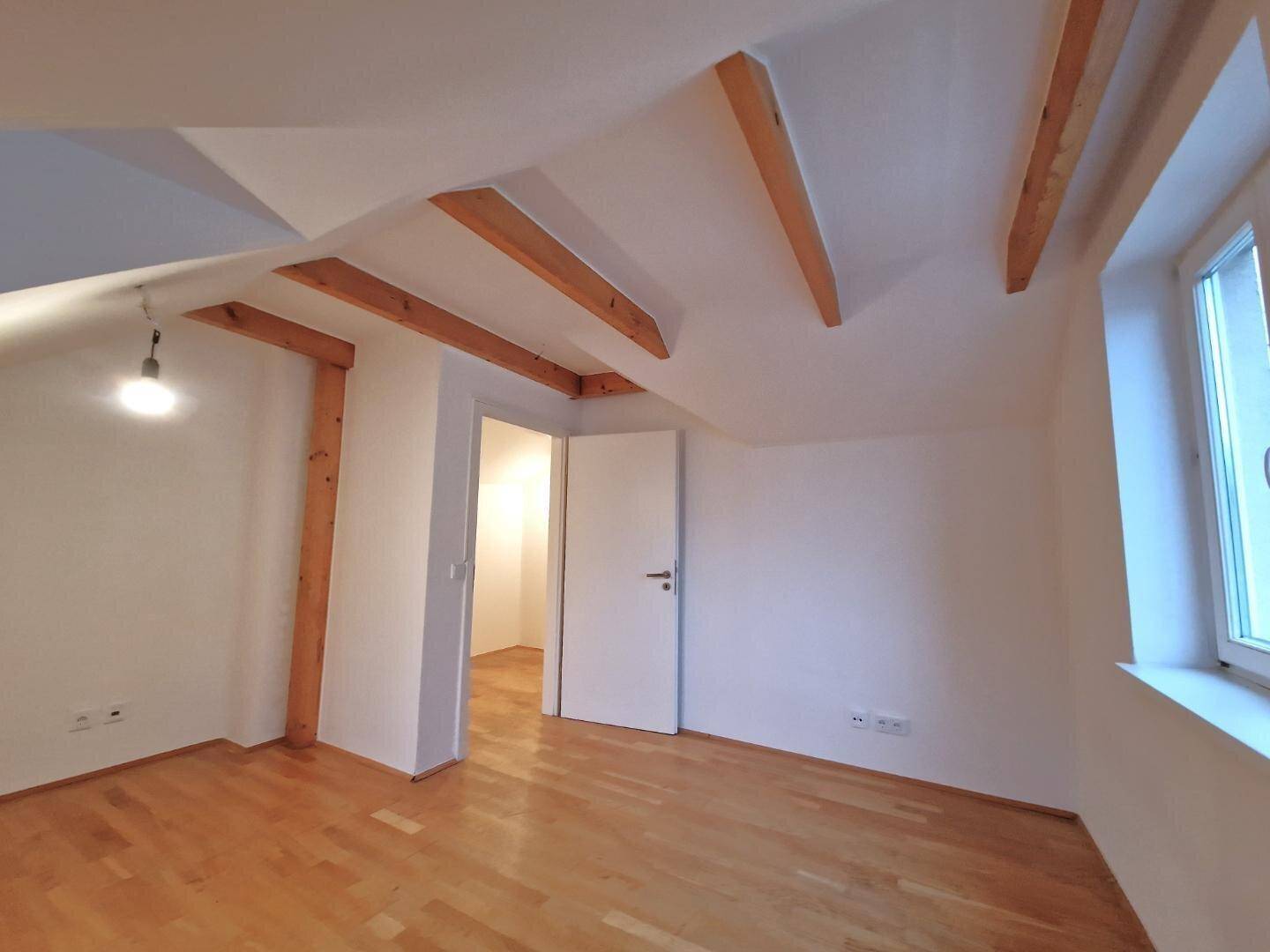Geräumiges, leeres Zimmer im Dachgeschoss mit Holzbalken und hellem Parkettboden.