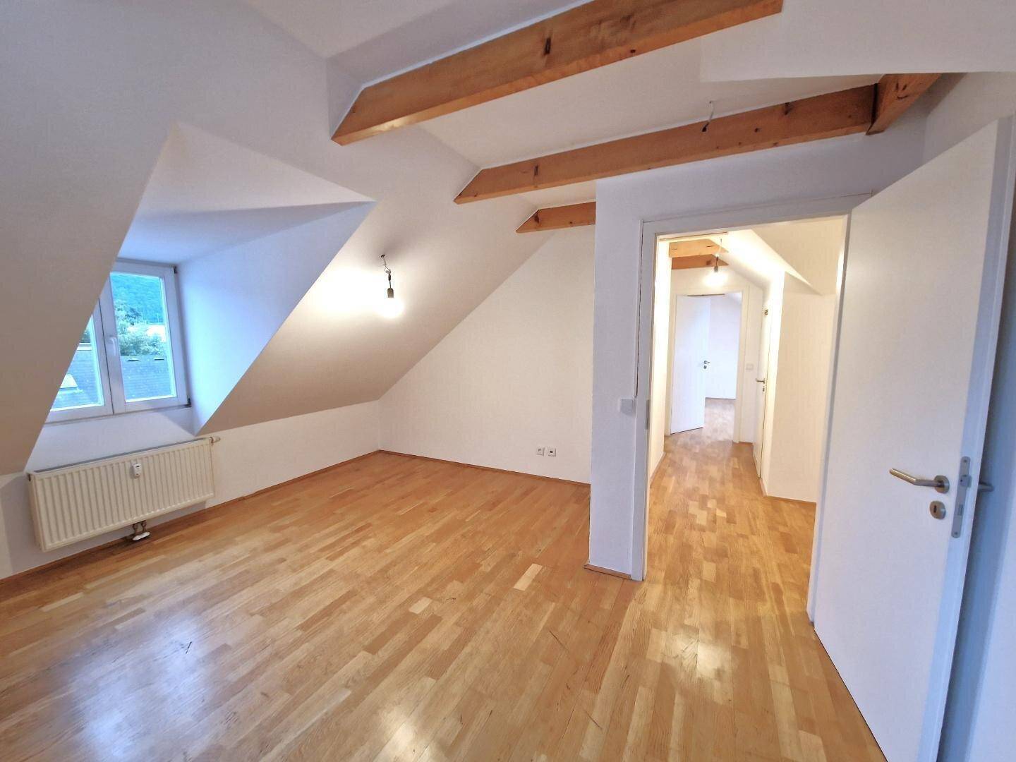 Helles, leeres Dachzimmer mit sichtbaren Holzbalken, Schrägdach und schönem Parkettboden.
