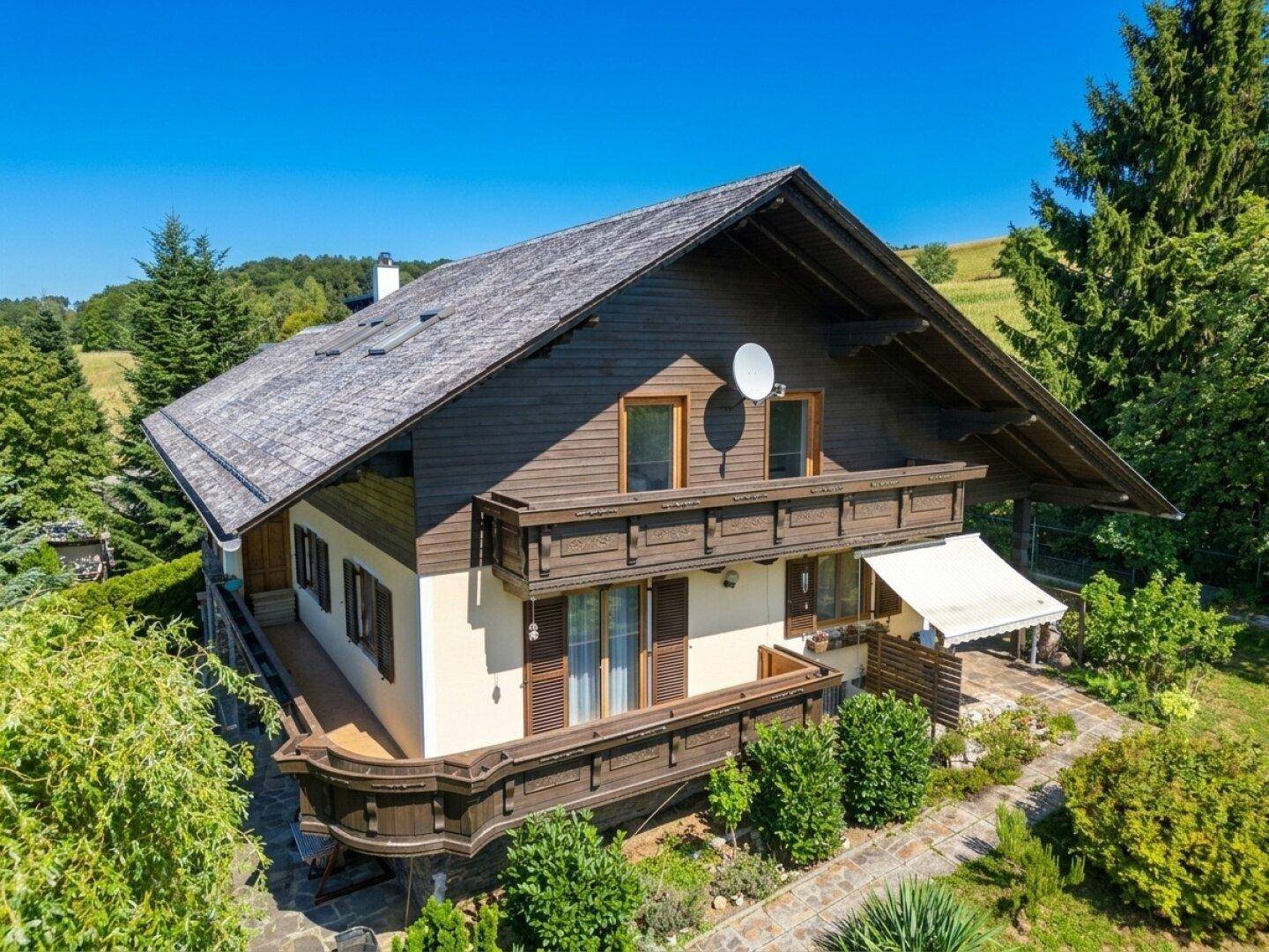 Traditionelles Landhaus mit Holzbalkonen und Satteldach, umgeben von grüner Natur unter blauem Himmel.