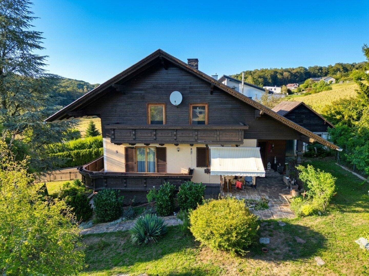 Traditionelles Landhaus mit Holzbalkonen und Satteldach, umgeben von grüner Natur unter blauem Himmel.