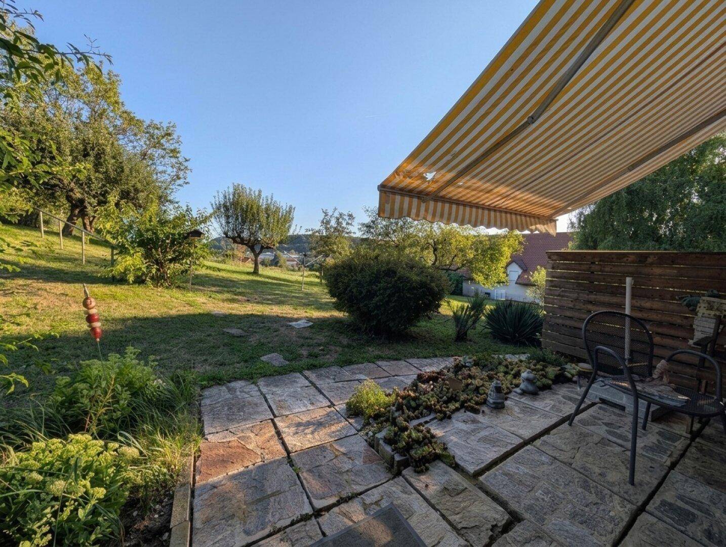 Weitläufiger Garten mit Rasenfläche, einer ausgefahrenen Markise und angrenzender Steinterrasse.