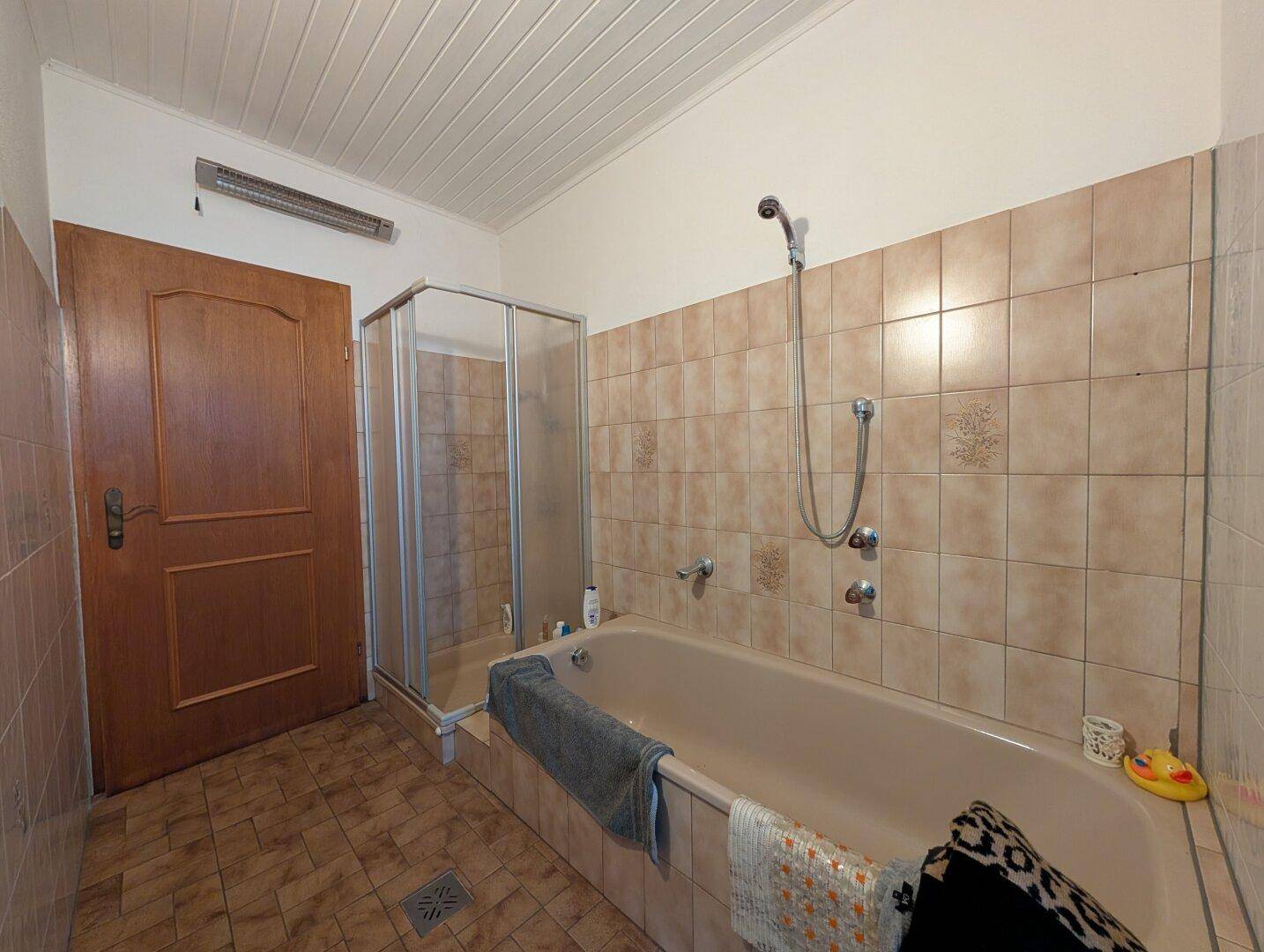 Badezimmer mit beigefarbenen Fliesen, Badewanne und Duschkabine, ausgestattet mit einem Heizstrahler.