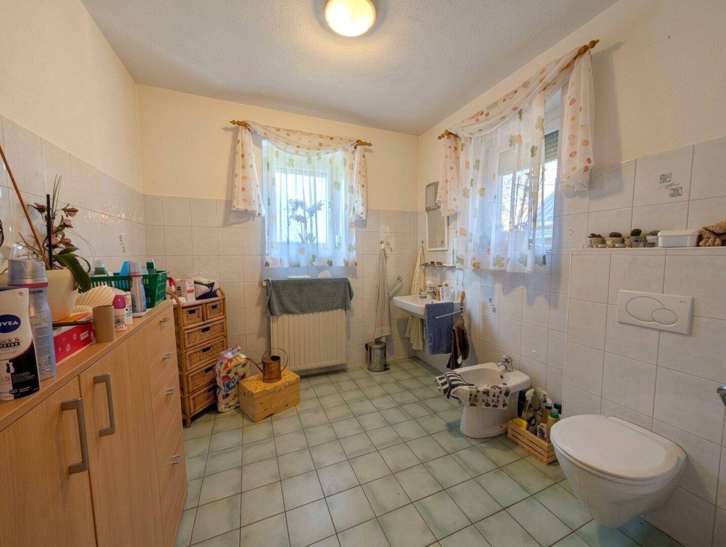 Geräumiges Badezimmer mit hellblauen Fliesen, Fenster, WC, Bidet und einem Waschbecken.