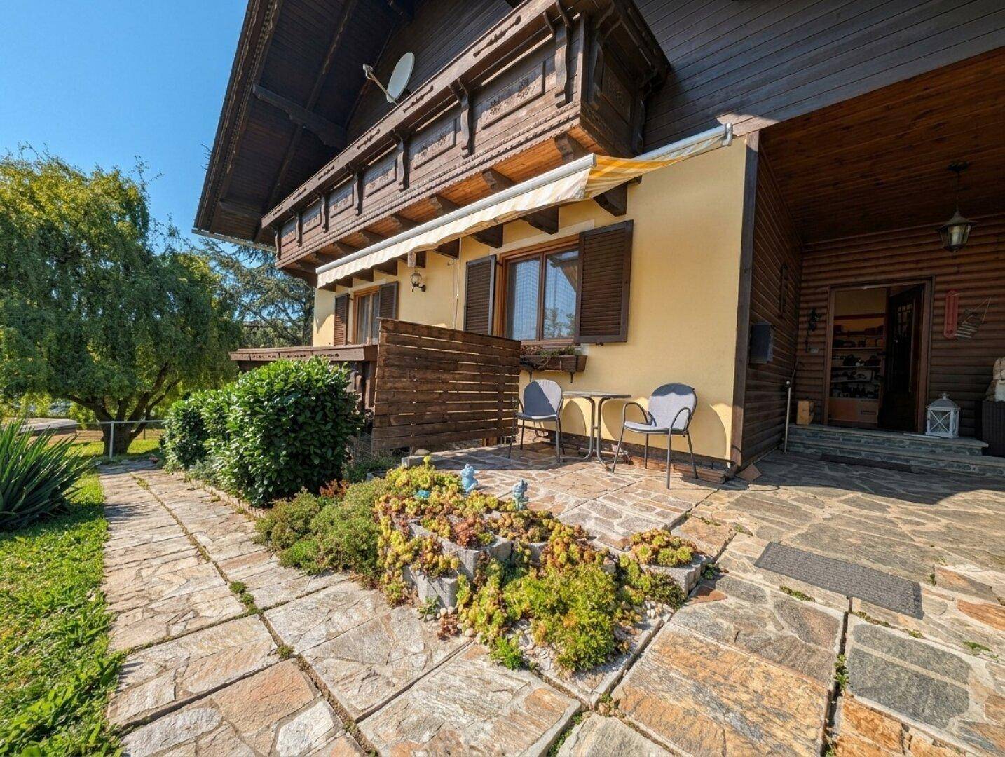 Sonniger Terrassenbereich mit Steinplatten, Gartenmöbeln und einer Markise, umgeben von Pflanzen.