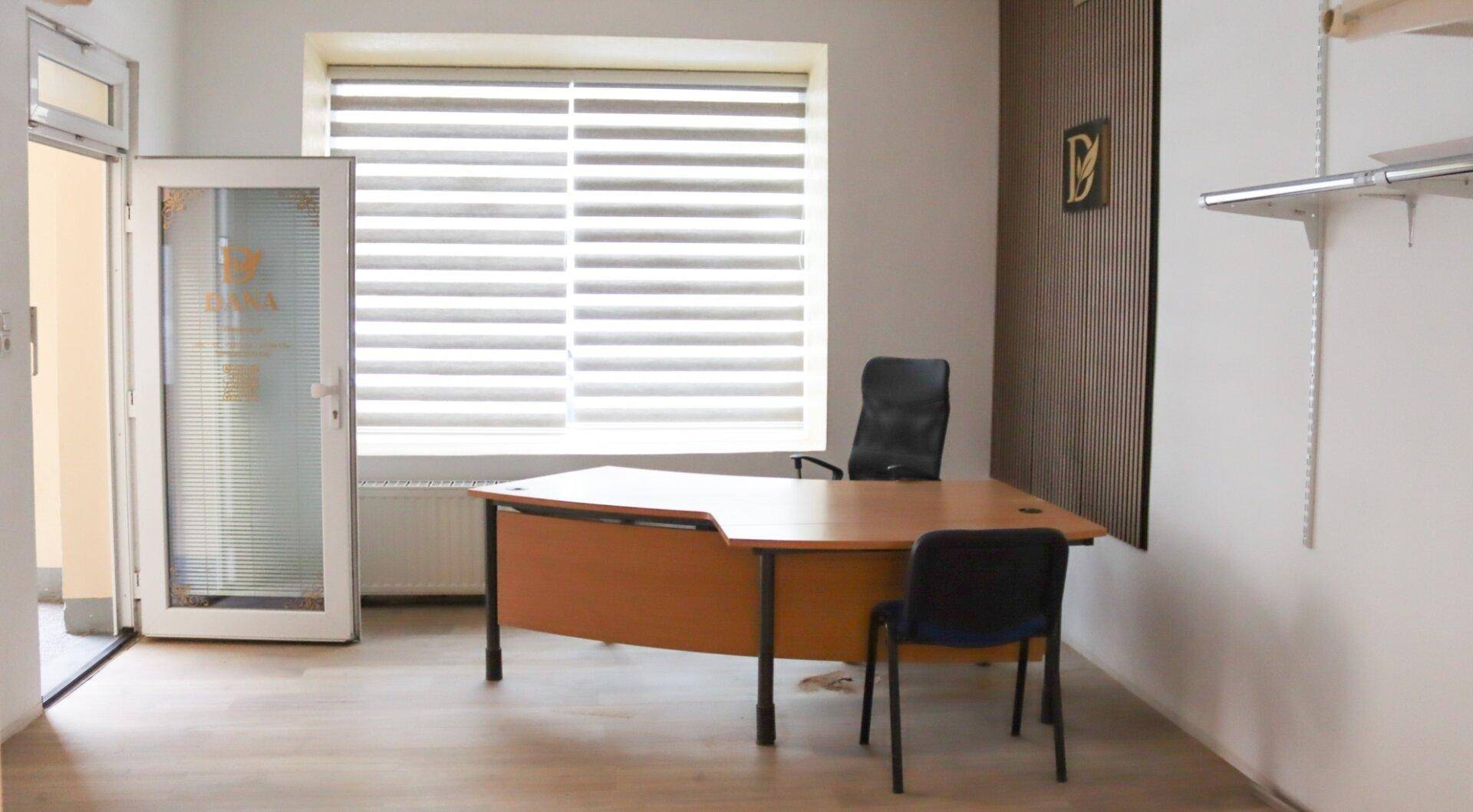 Büroraum mit einem Schreibtisch, Bürostuhl und einem Fenster mit Jalousien, das Tageslicht hereinlässt.