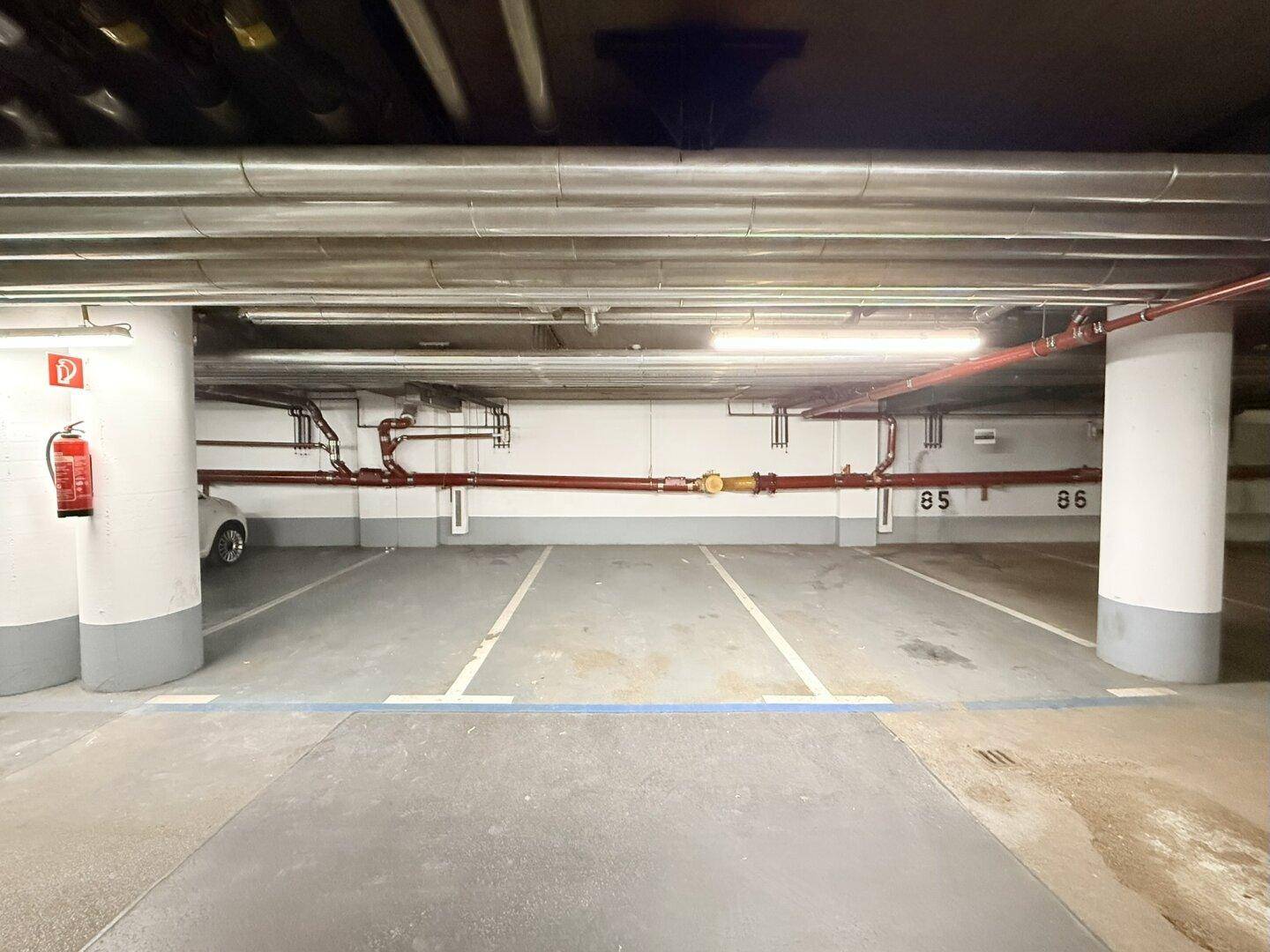 Zwei Parkplätze in einer Tiefgarage mit sichtbaren Versorgungsleitungen und einem Feuerlöscher an der Wand.