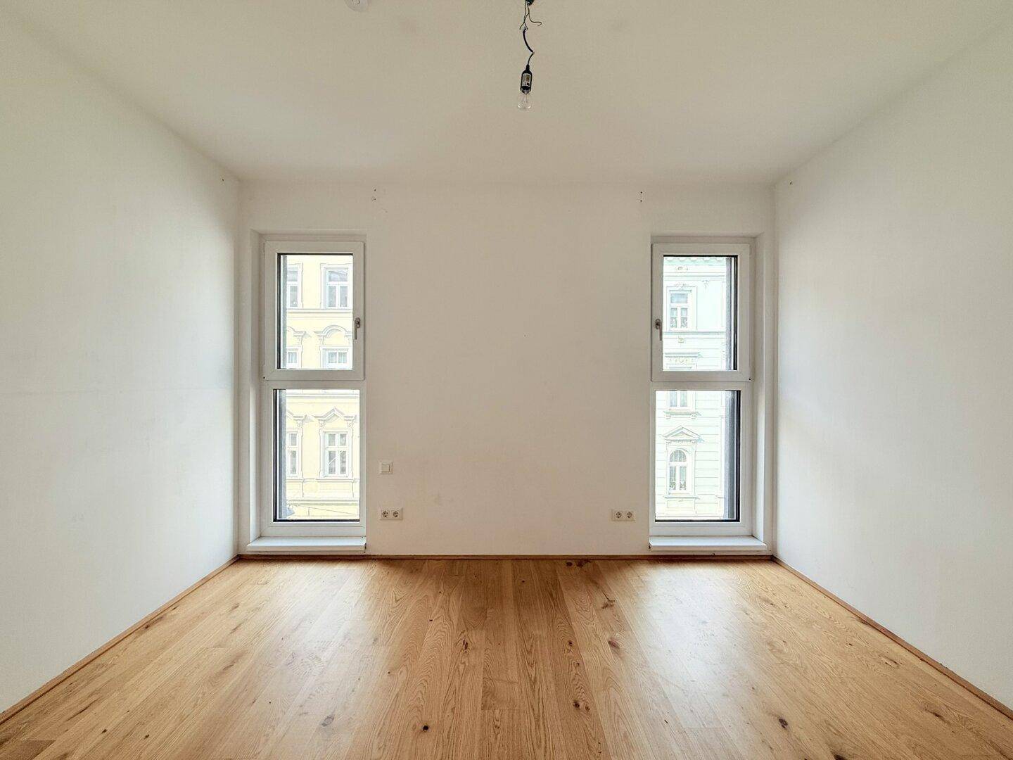Leeres Zimmer mit zwei hohen Fenstern und Holzboden, ideal für individuelle Gestaltung.