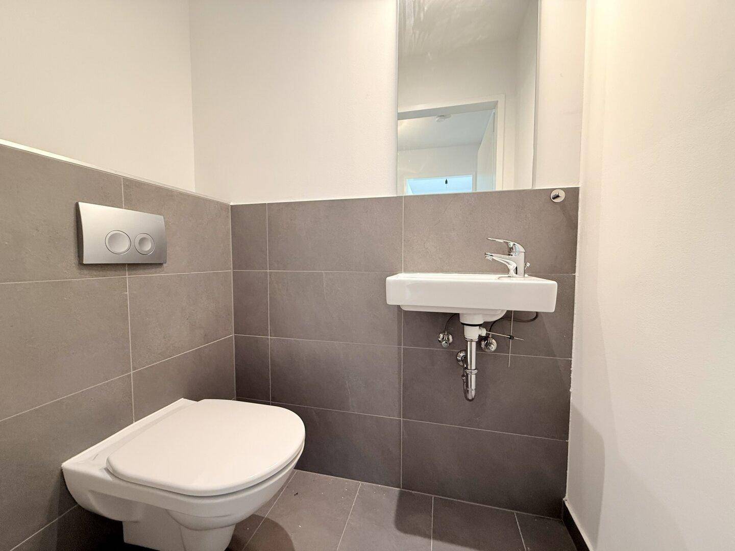 Separates Gäste-WC mit Wand-WC, kleinem Waschbecken und modernen grauen Fliesen.