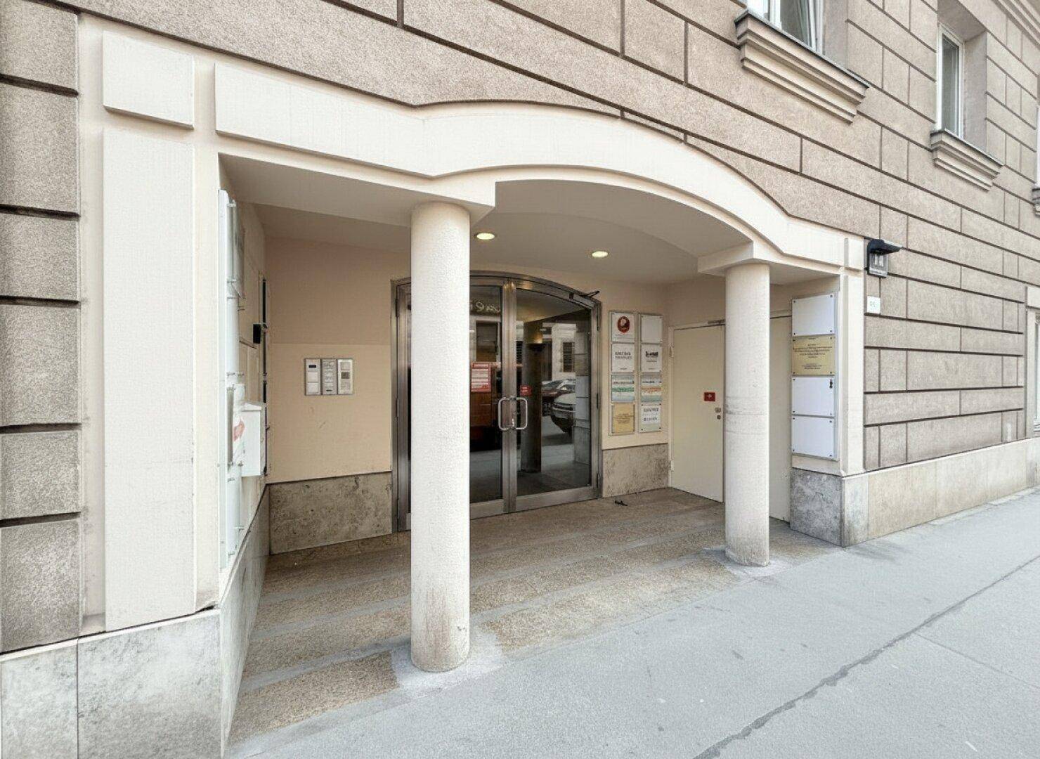 Gebäudeeingang mit dekorativen Säulen, einem Vordach und einer Glastür, umgeben von Hinweisschildern.