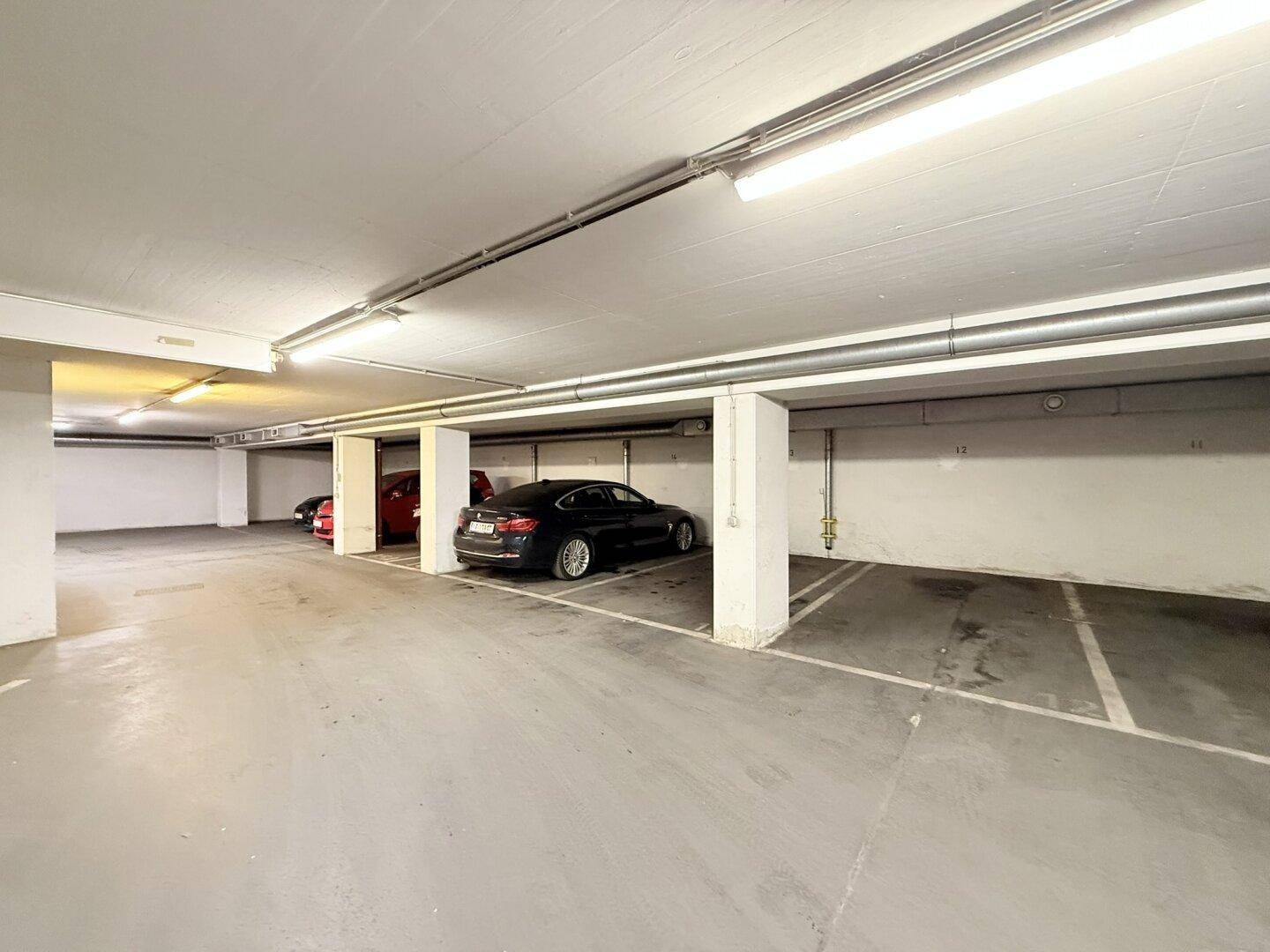 Tiefgarage mit mehreren Stellplätzen und sichtbaren technischen Installationen an der Decke und Wänden.