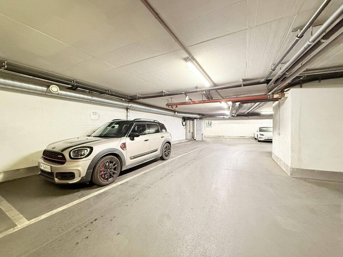 Geräumige Tiefgarage mit hellen Wänden und Decken, sowie sichtbaren Rohrleitungen und Beleuchtung.