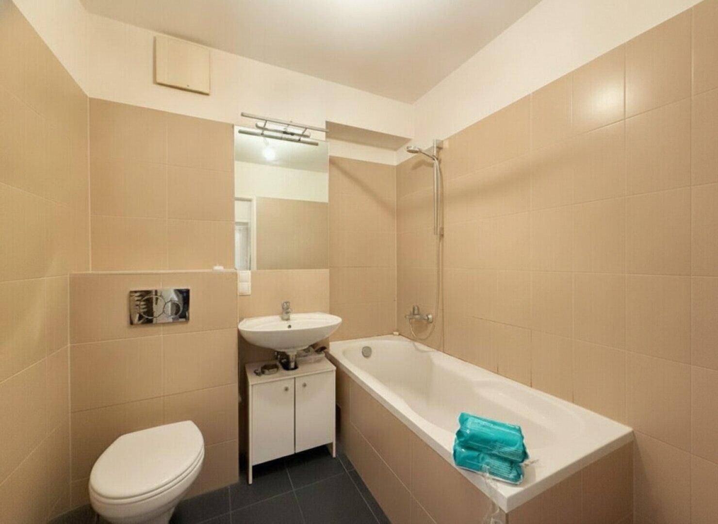 Helles Badezimmer mit beigefarbenen Fliesen, Badewanne mit Duschfunktion, WC und Einzelwaschbecken.