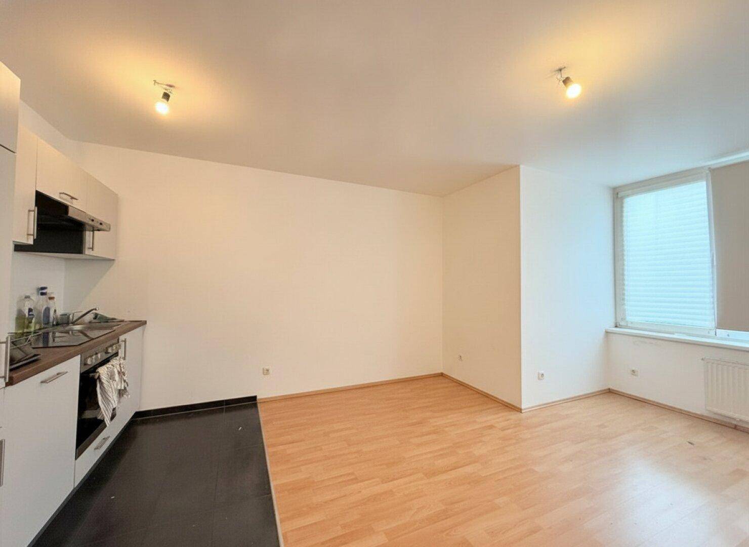 Offener Wohnbereich mit integrierter Küchenzeile, Parkettboden und viel Tageslicht durch das Fenster.