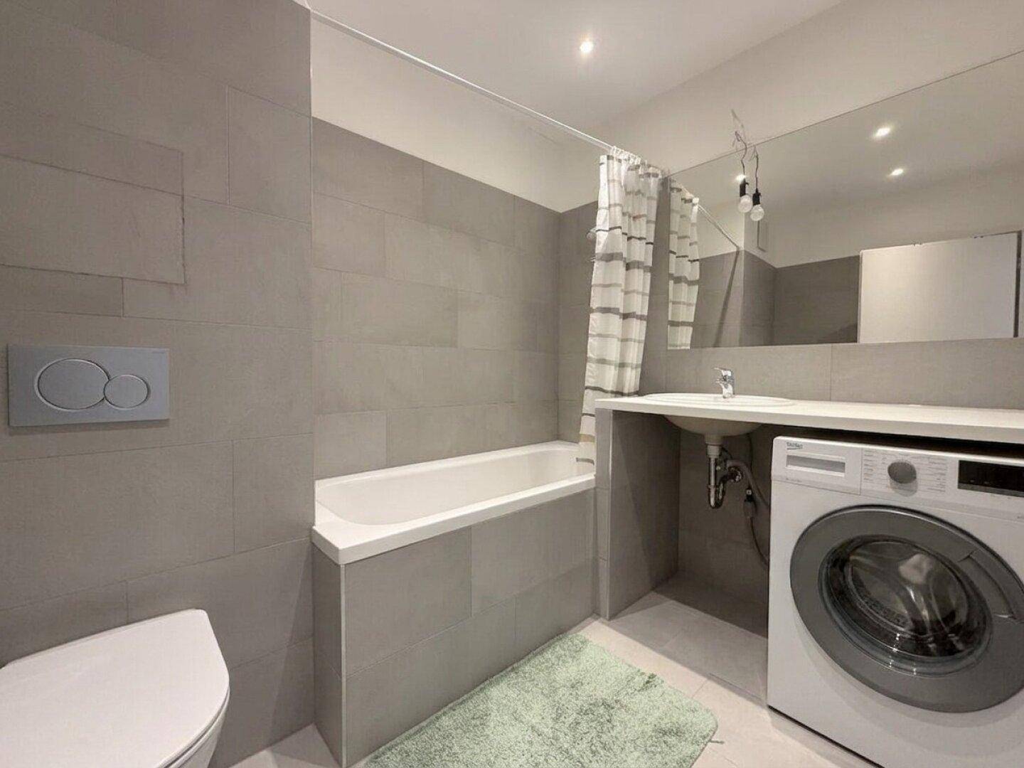 Modernes Badezimmer mit grauen Fliesen, Badewanne und Waschmaschinenanschluss, funktional und hell.
