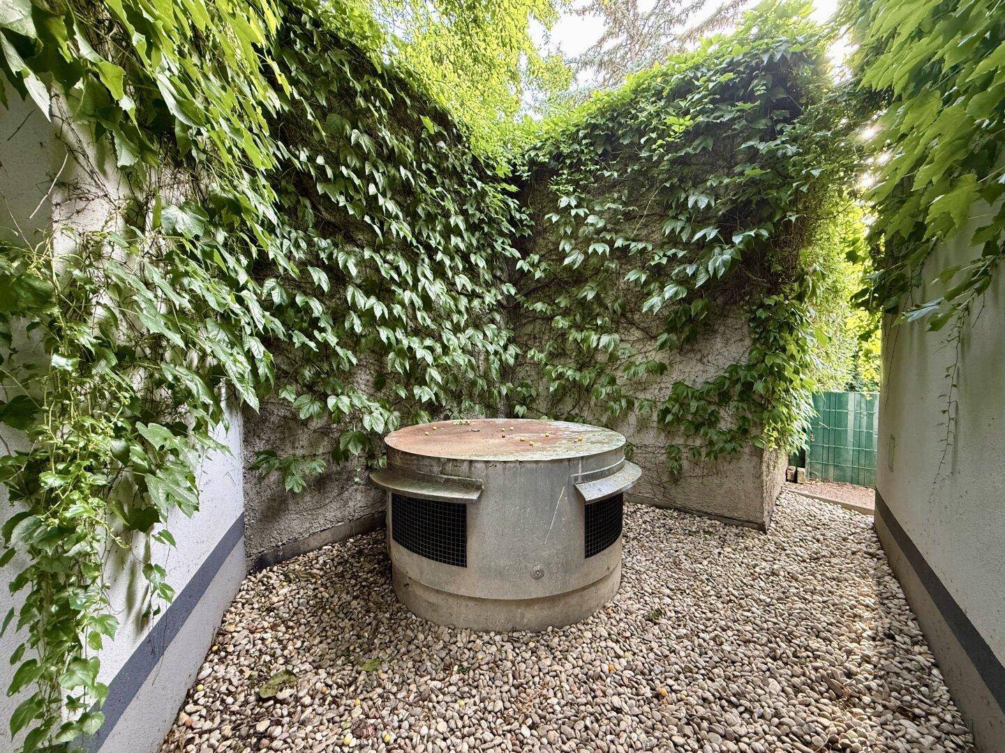 Umschlossener Außenbereich mit Kiesboden und Efeu bewachsenen Wänden, mit einer runden Betonstruktur in der Mitte.