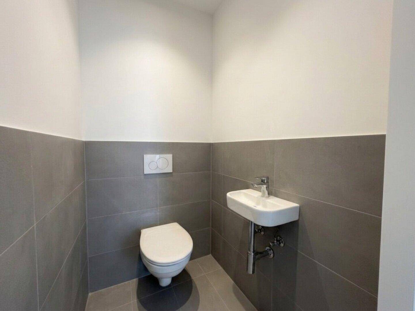 Funktionelles Gäste-WC mit wandhängender Toilette, kleinem Waschbecken und grauen Wandfliesen.