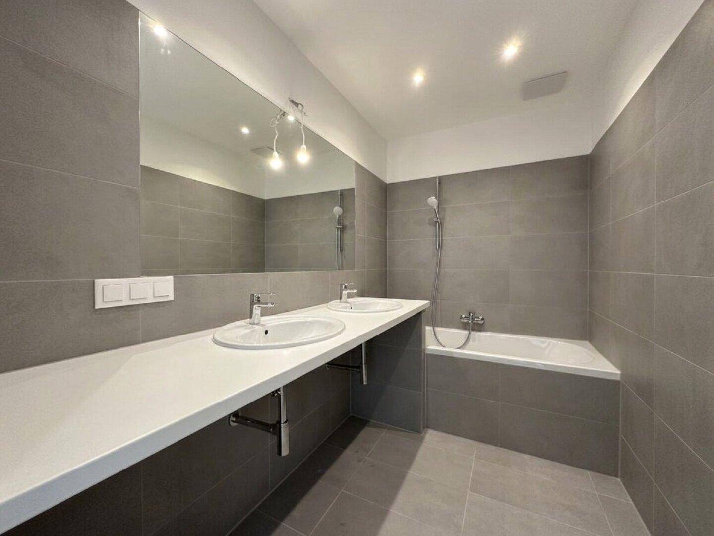 Modernes Badezimmer mit Doppelwaschbecken, Badewanne und großen grauen Fliesen.