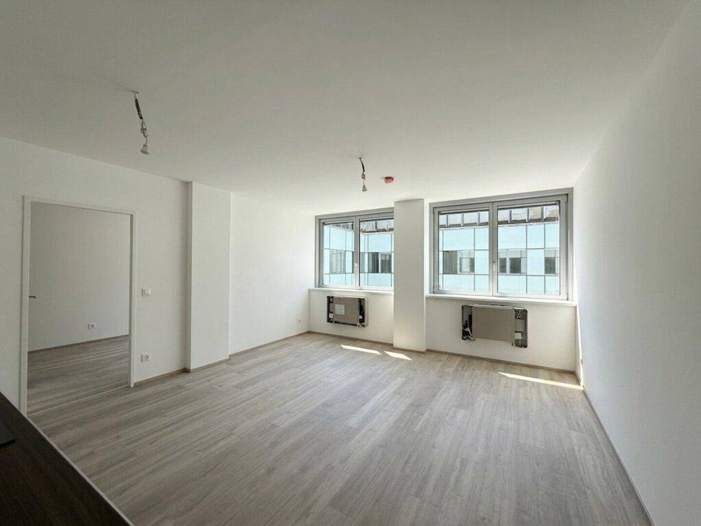 Geräumiges Wohnzimmer mit hellem Laminatboden und großen Fenstern, die viel Tageslicht hereinlassen.