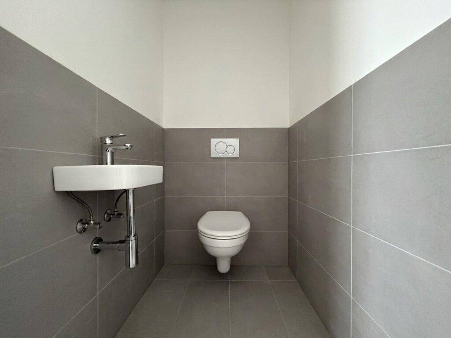 Modernes Gäste-WC mit grauen Fliesen, wandhängendem WC und kleinem Waschbecken.