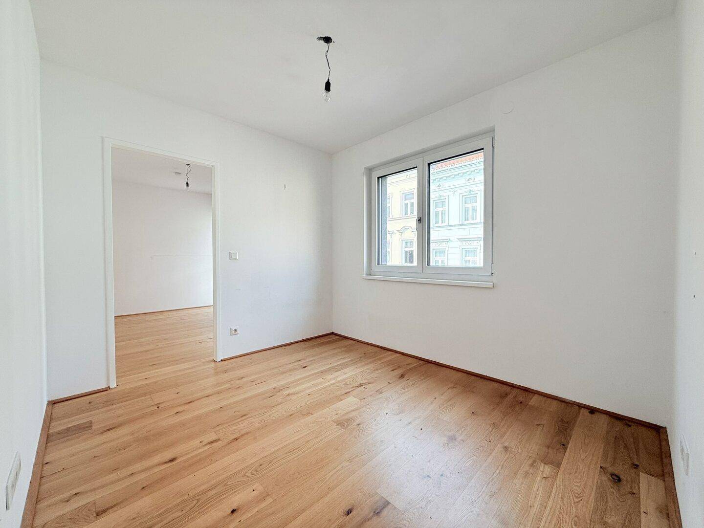Leeres Zimmer mit Holzboden und Fenster, das einen Blick auf die gegenüberliegende Fassade bietet.