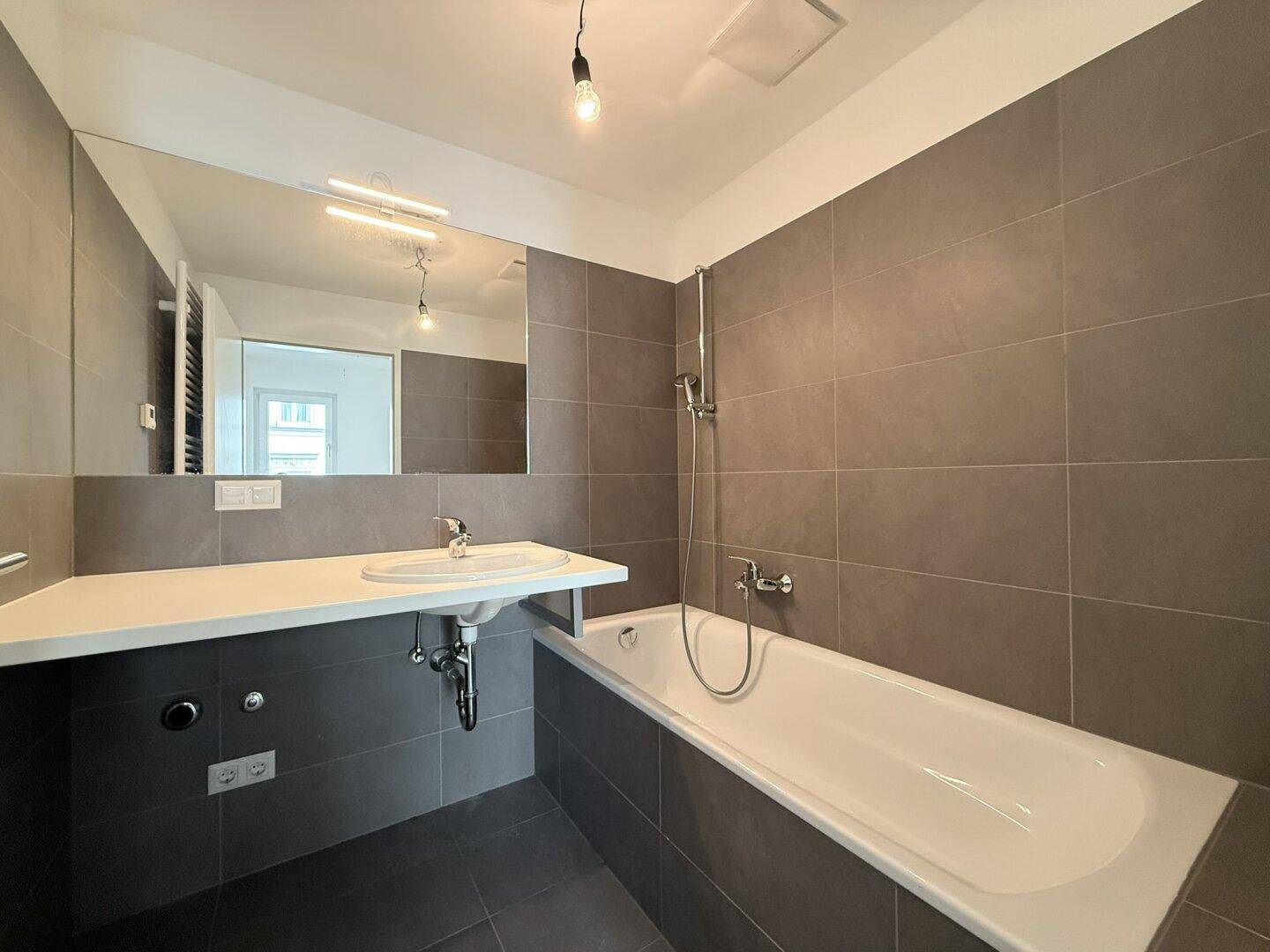 Modernes Badezimmer mit grauen Fliesen, Badewanne und großem Spiegel über dem Waschbecken.